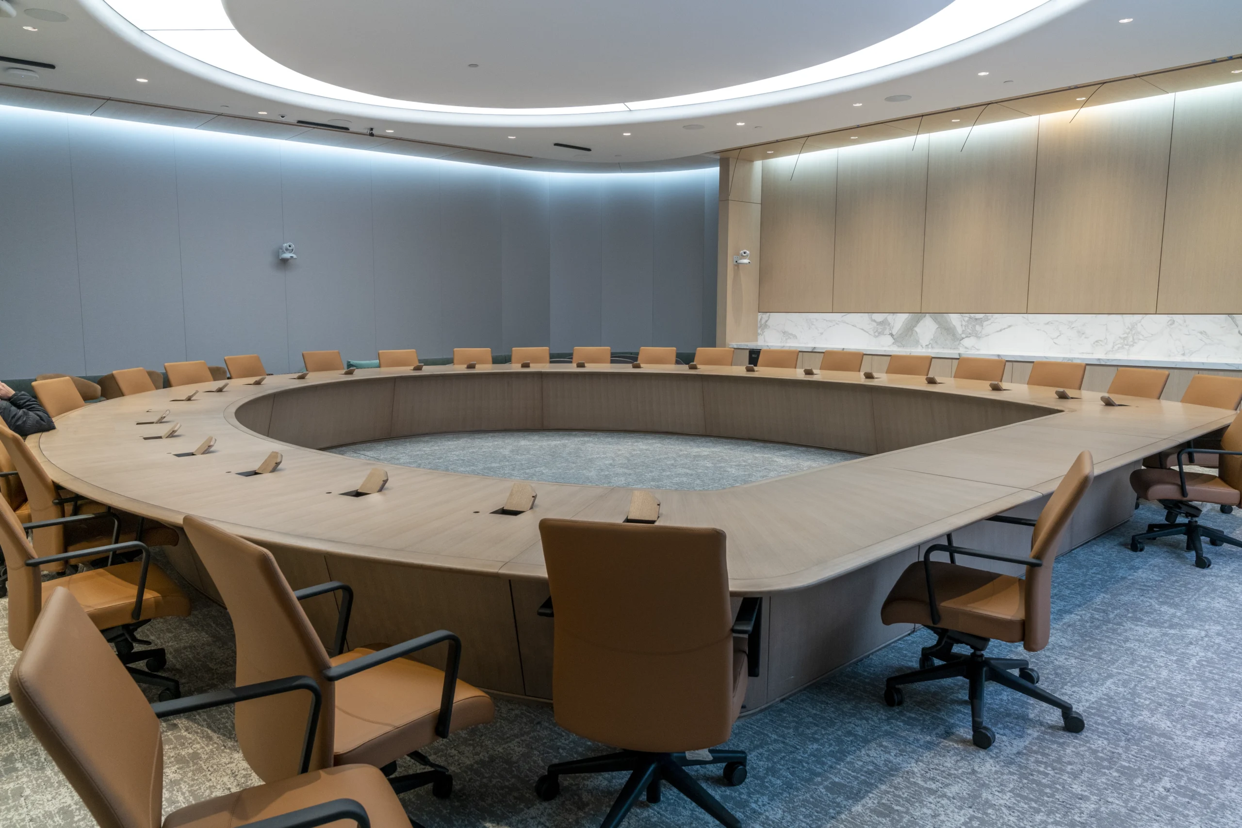 Modern corporate boardroom with a large bleached walnut table surrounded by tan chairs, equipped with microphones and notepads, featuring a minimalist design with wood panels and subtle lighting.