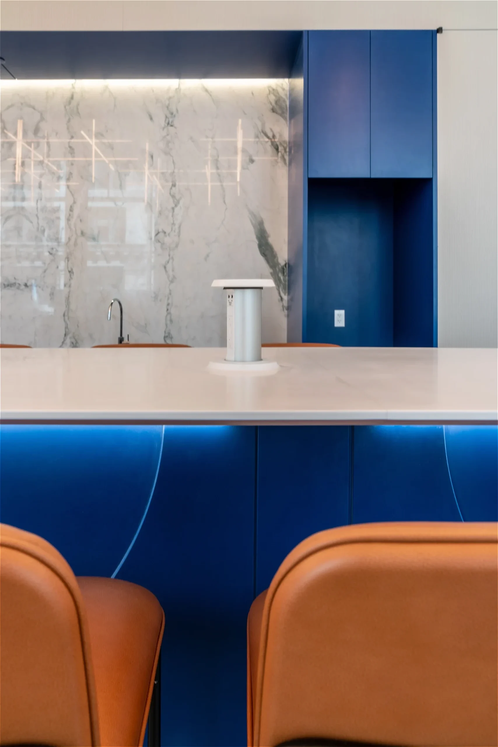 Modern kitchen interior featuring a marble backsplash, blue cabinets, white Corian countertop with a minimalistic white appliance, and orange chairs at the coffee bar.