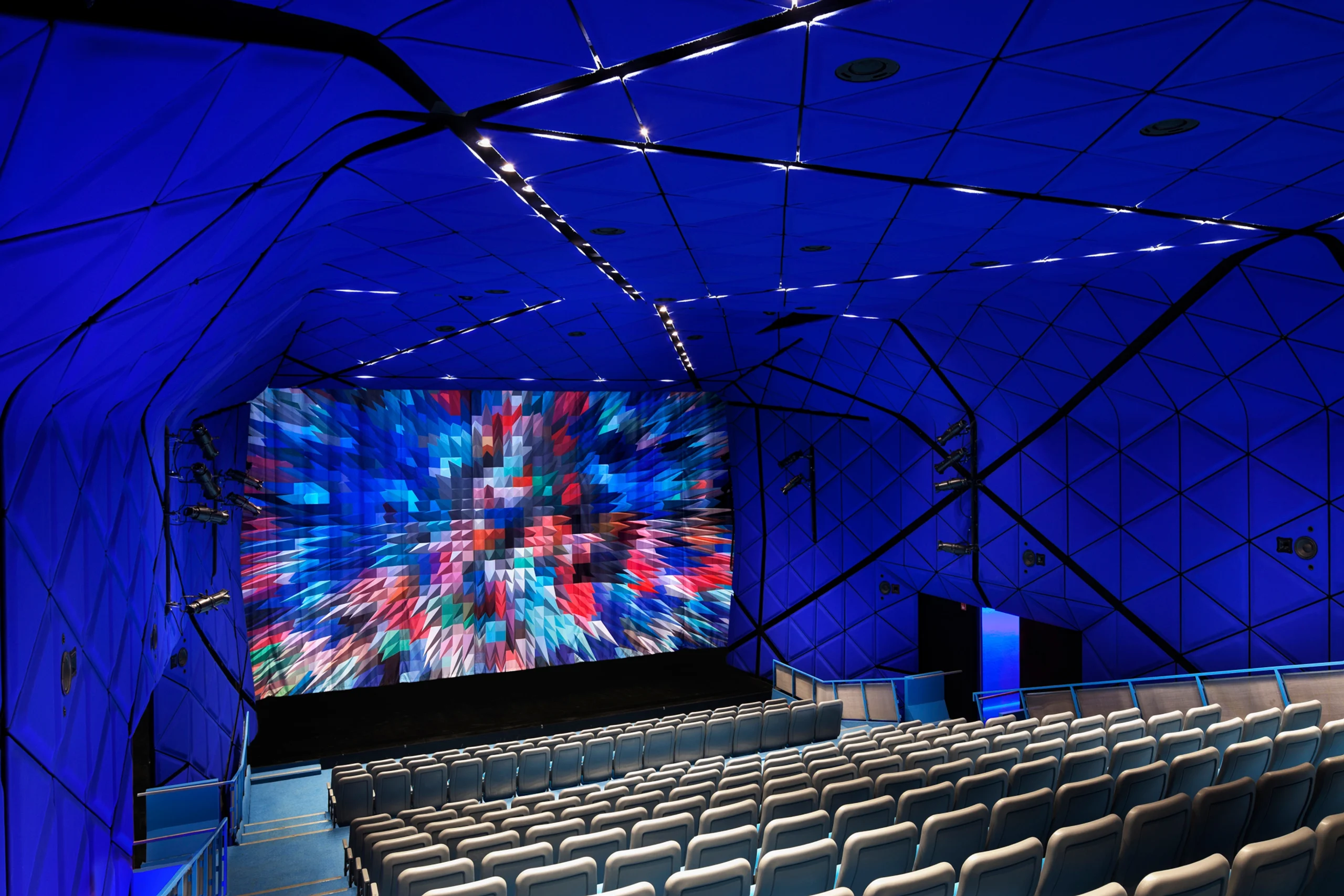 Modern cinema hall with blue geometric panel walls, star-like ceiling lights, and filled rows of gray seats facing a large screen displaying vibrant graphics.