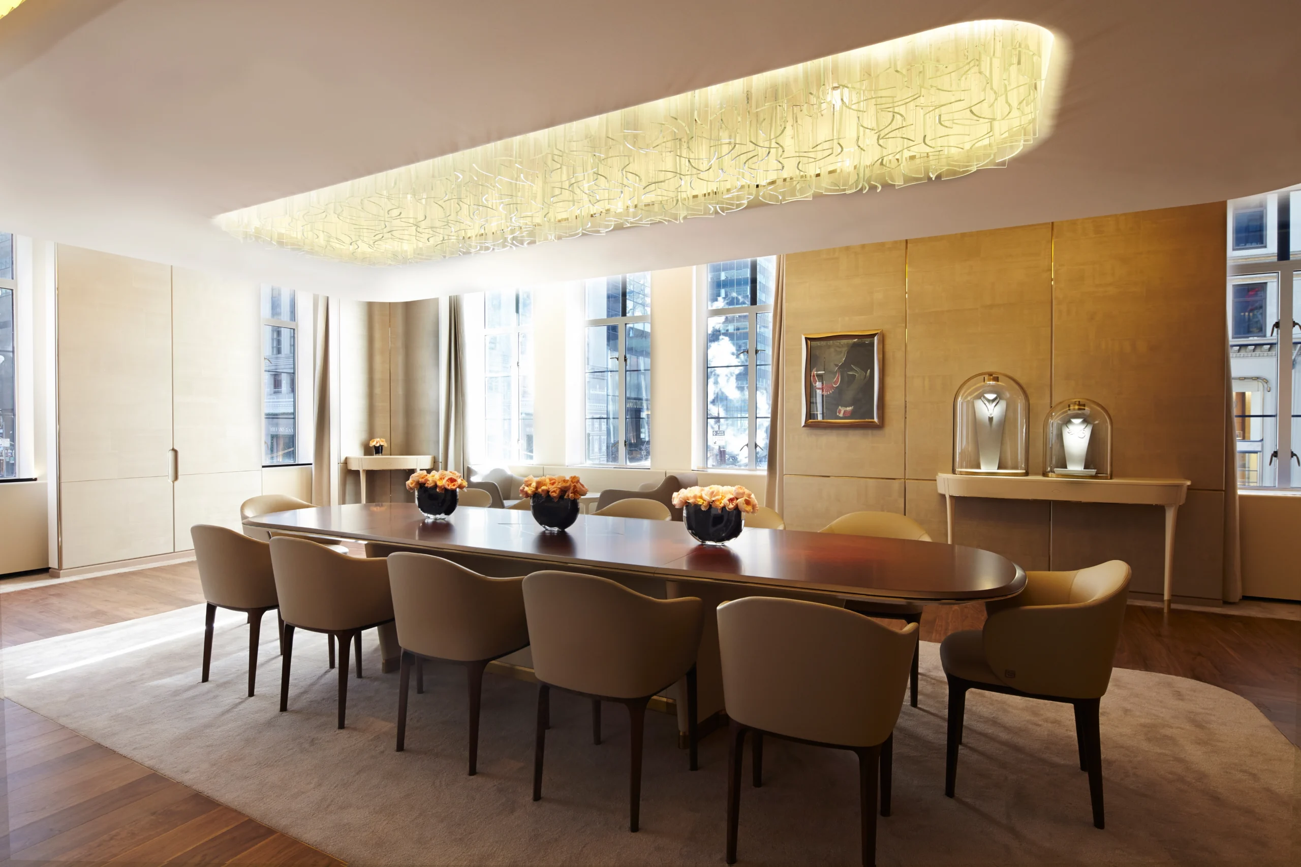 Elegant dining room with a long oval table, tan chairs, and a large illuminated oval ceiling fixture, surrounded by modern decor and large windows.